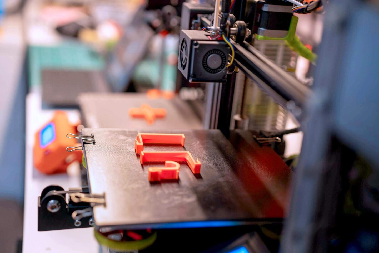 Ein 3D gedrucktes Bauteil liegt auf einer Fläche vor einem 3D Drucker